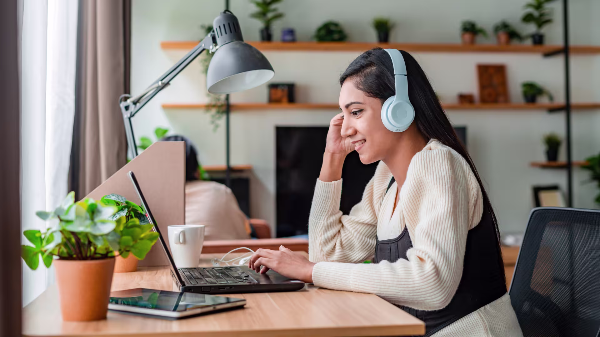 Person wearing white sweater and headphones works on laptop at desk with houseplants in bright modern office setting