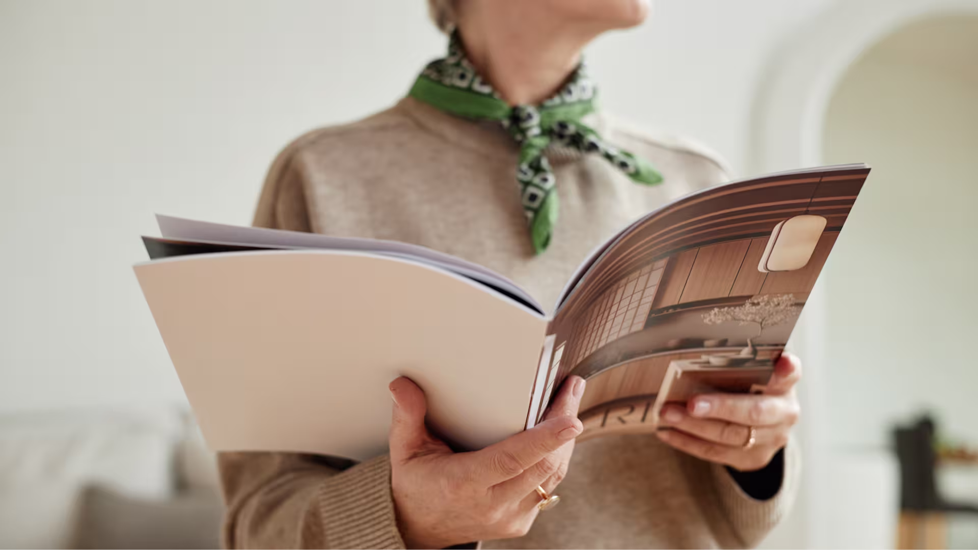 A woman in a beige sweater and green scarf holds an open interior design magazine showing a wooden room.