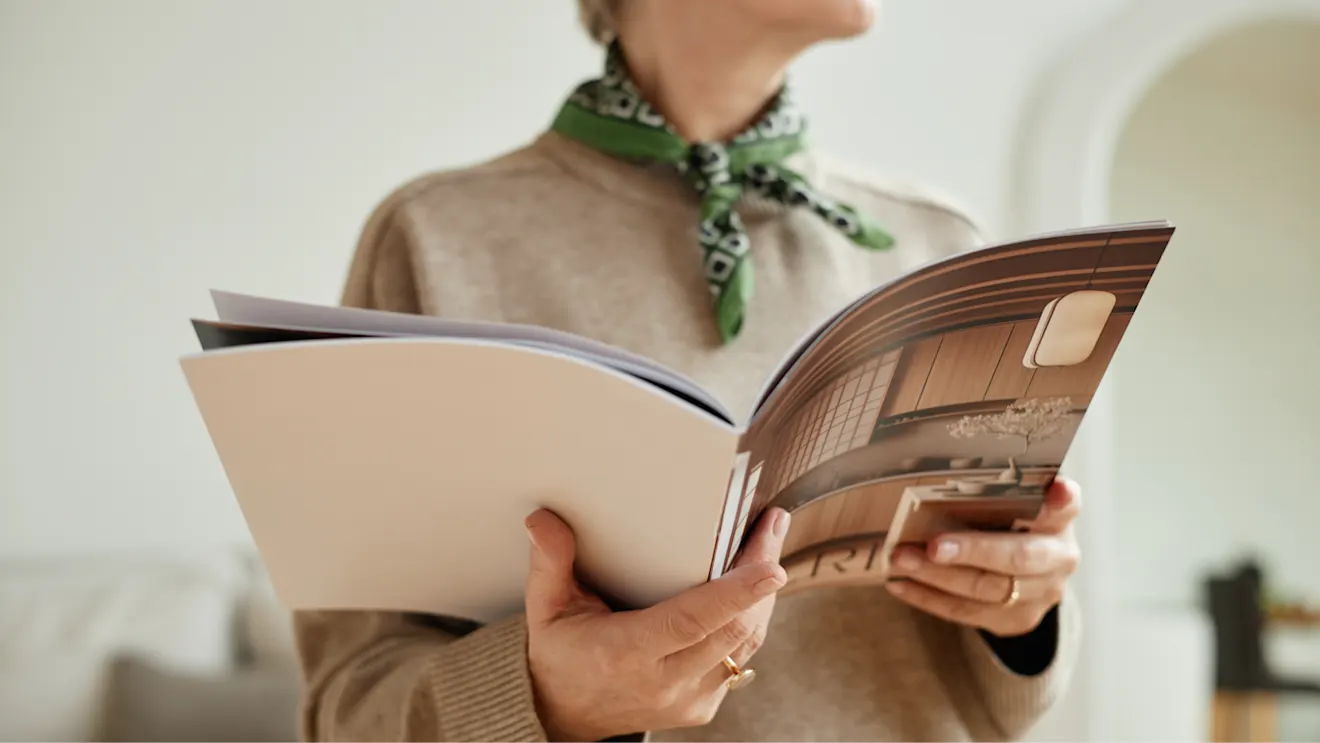 A woman in a beige sweater and green scarf holds an open interior design magazine showing a wooden room.