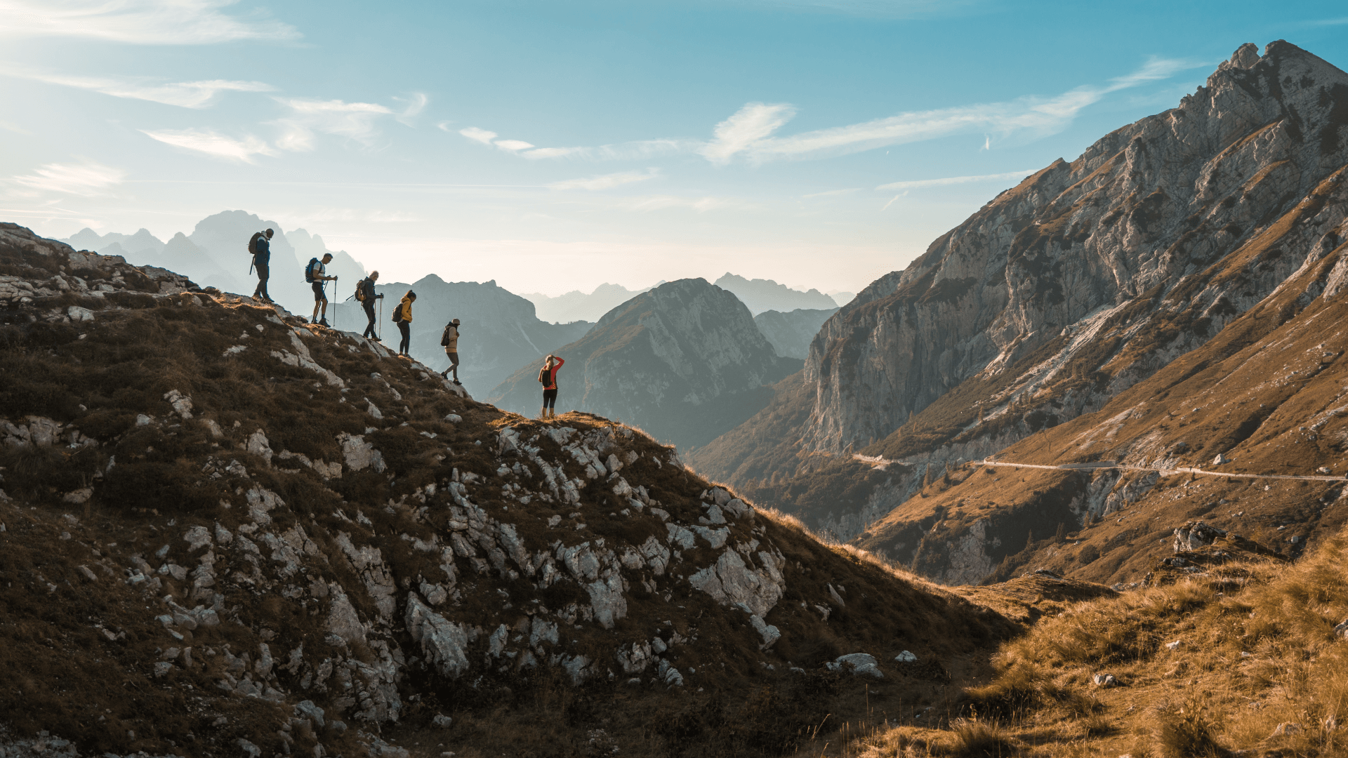 Hikers traverse a rocky mountain ridge at sunset, silhouetted against dramatic alpine peaks and a winding valley road below