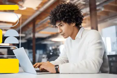 Young man with curly hair wearing white shirt working intently on laptop in modern cafe setting with warm lighting.