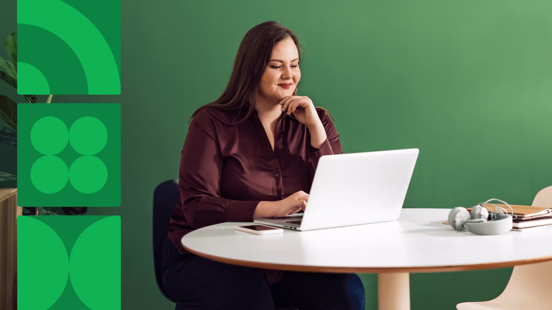 A woman in a burgundy blouse smiles while working on a laptop at a white table against a green wall.