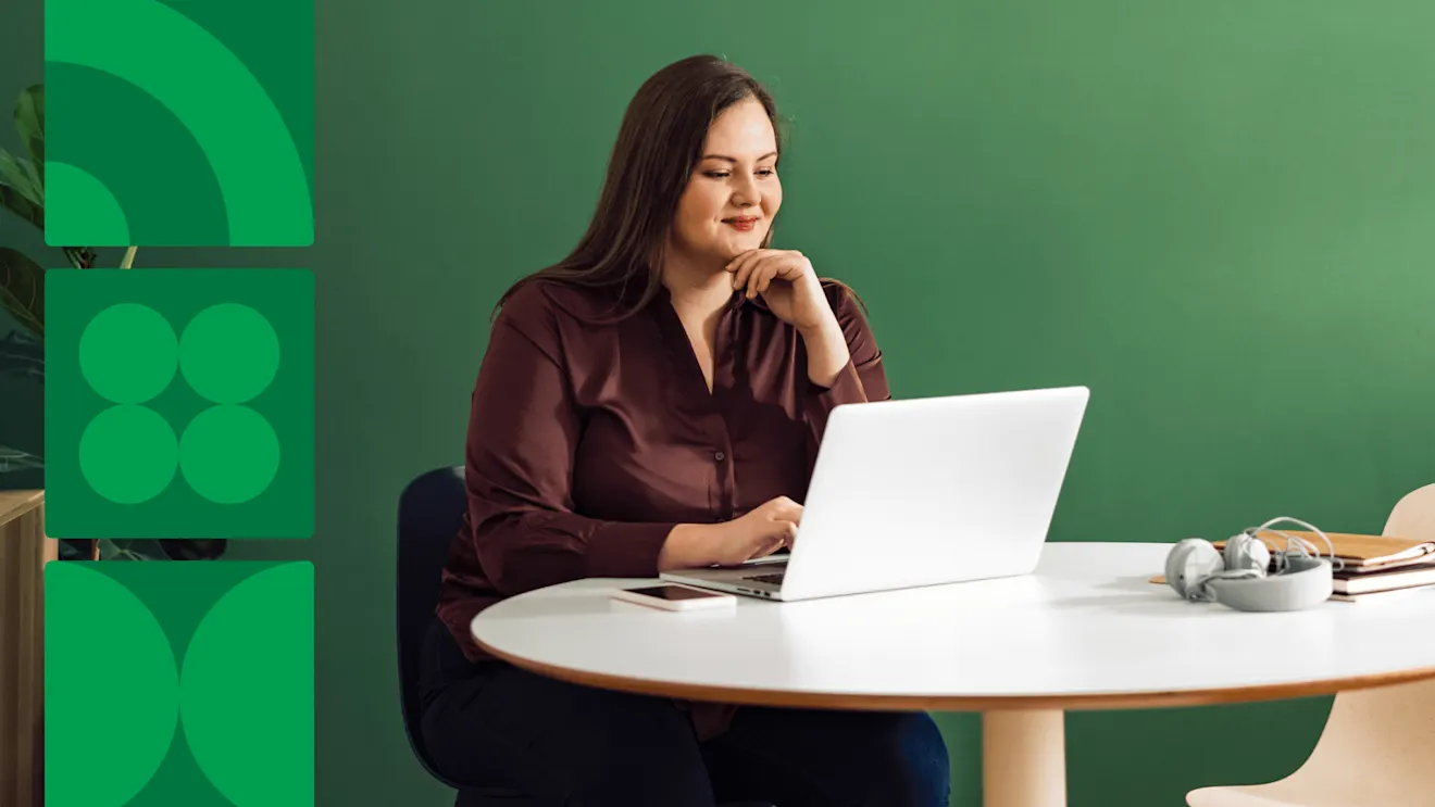 A woman in a burgundy blouse smiles while working on a laptop at a white table against a green wall.