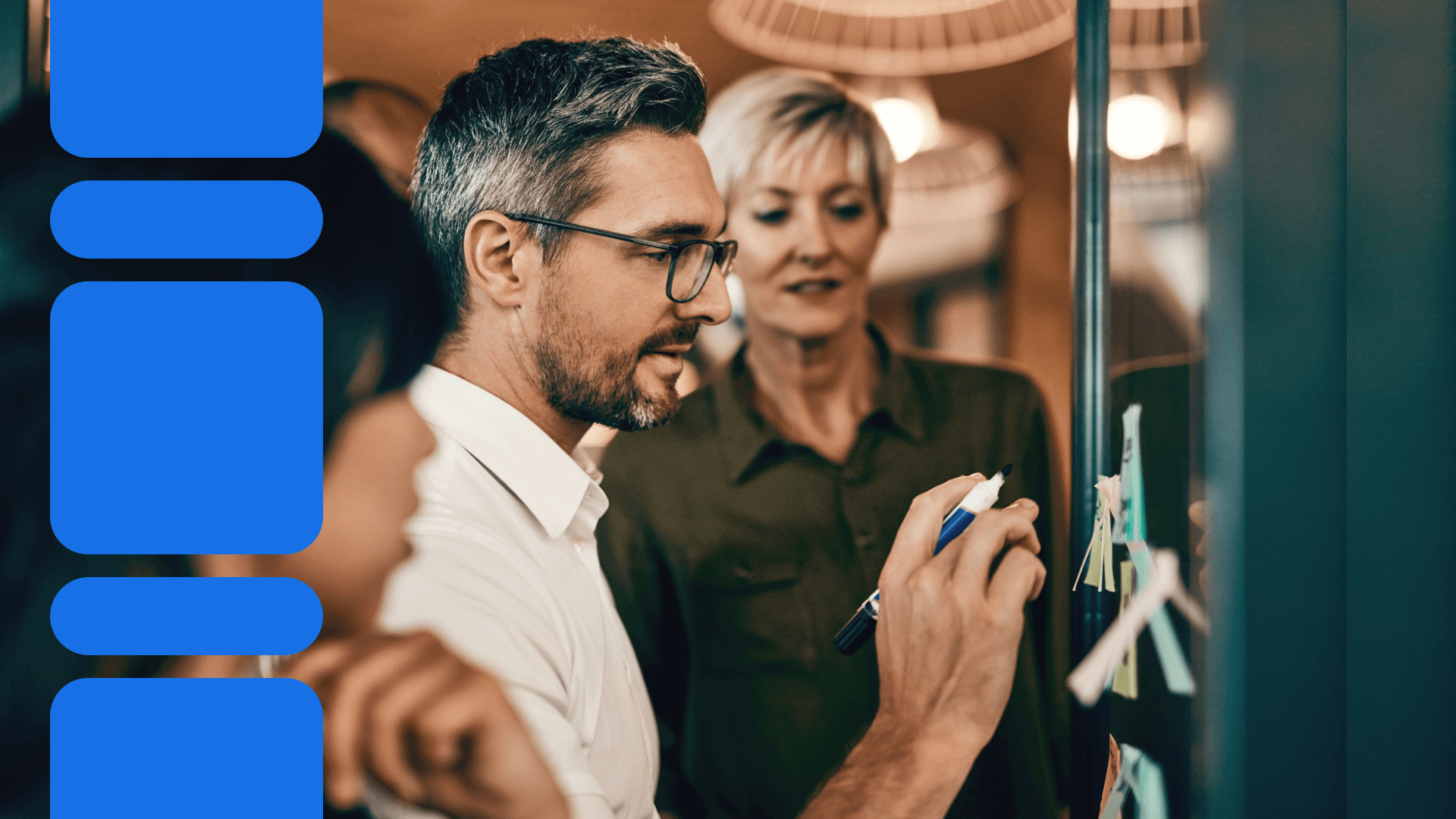 Two business professionals collaborating at a glass board, one writing with marker while the other observes thoughtfully