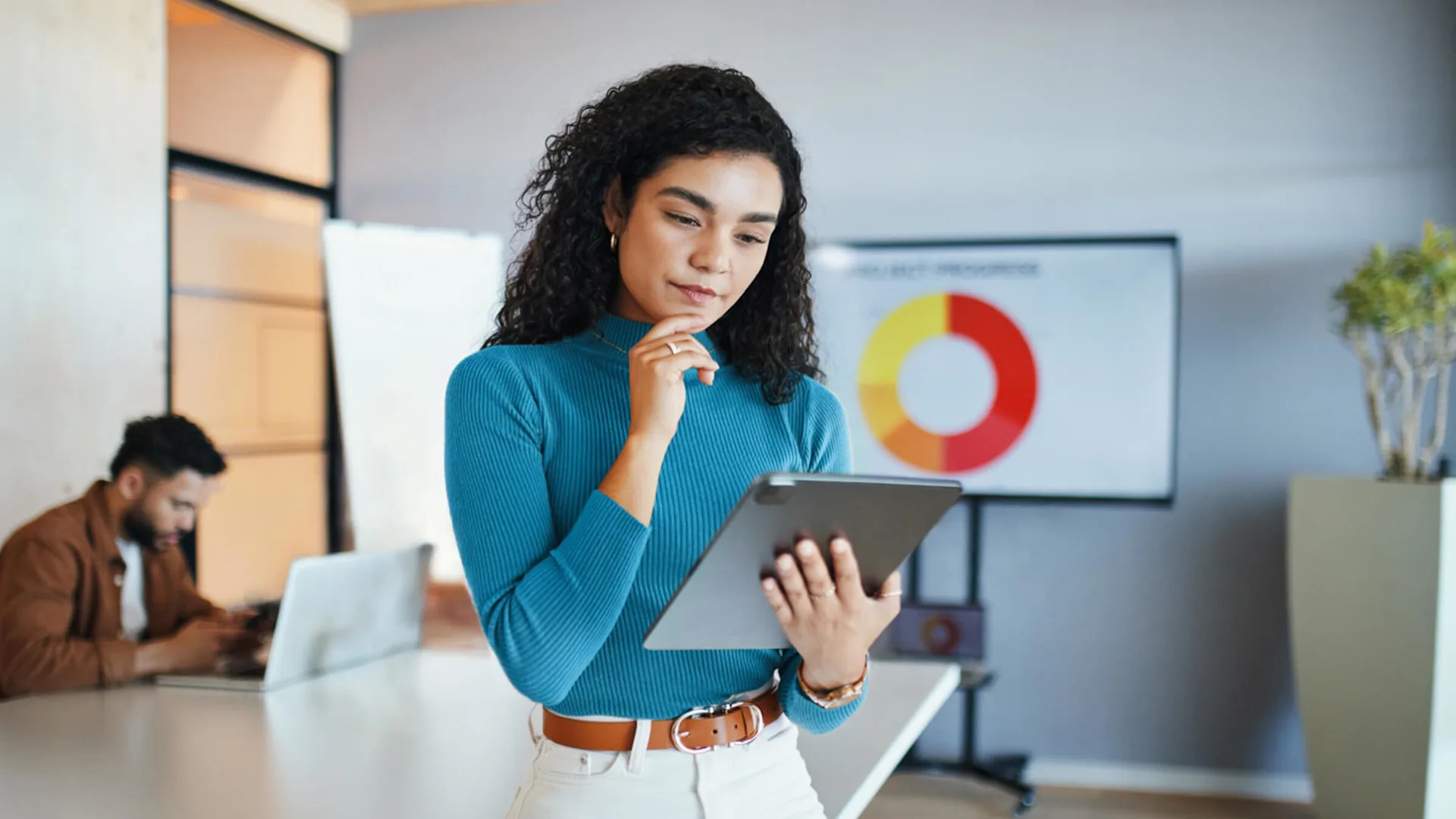 Professional in blue turtleneck reviews documents in modern office with colorful circular wall art and colleague in background