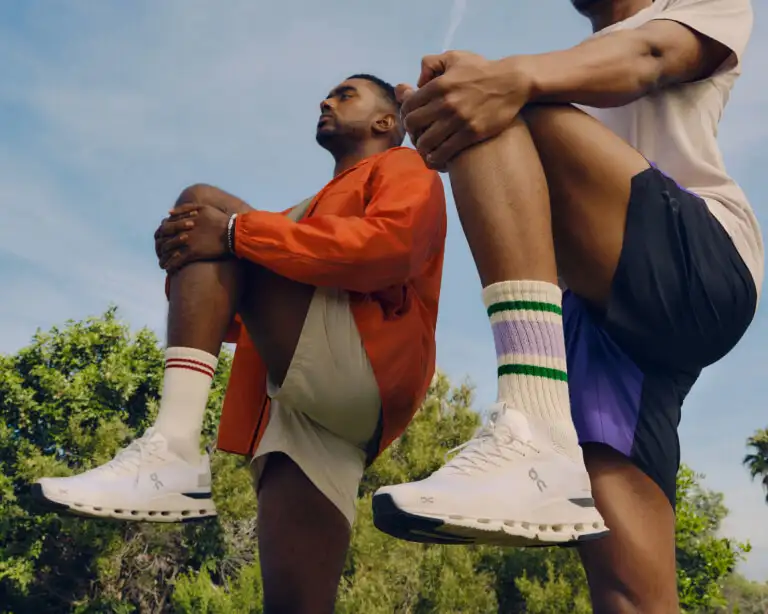 A pair of athletes stretch, wearing On running shoes.
