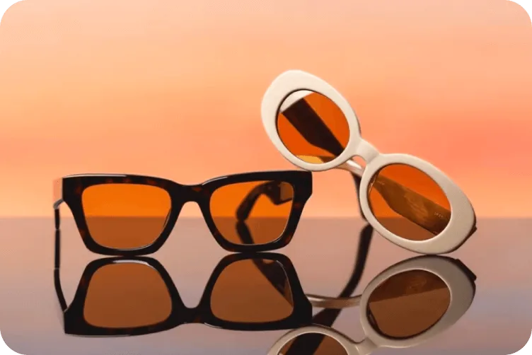 Two pairs of sunglasses with amber lenses - one black framed and one white framed - on reflective surface against orange background