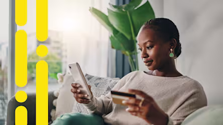 Woman sitting on a sofa holding a phone in one hand and a credit card in the other, with yellow graphic shapes along the left edge.