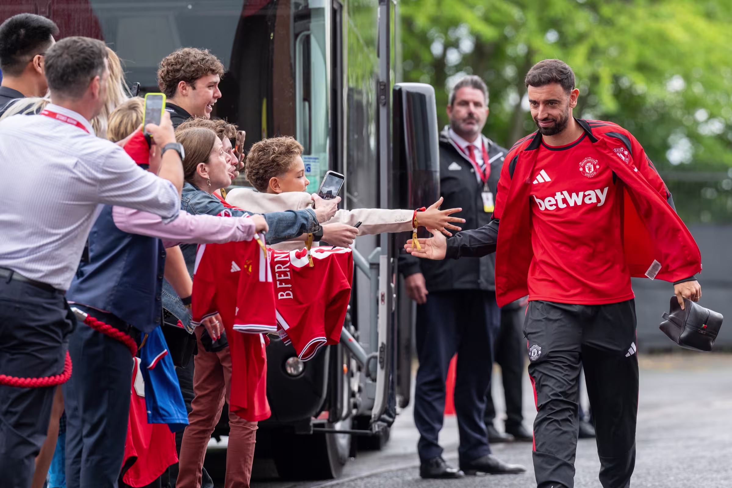 A Manchester United player in a red training top reaches out to shake hands with supporters behind a barrier as he walks from the team coach.