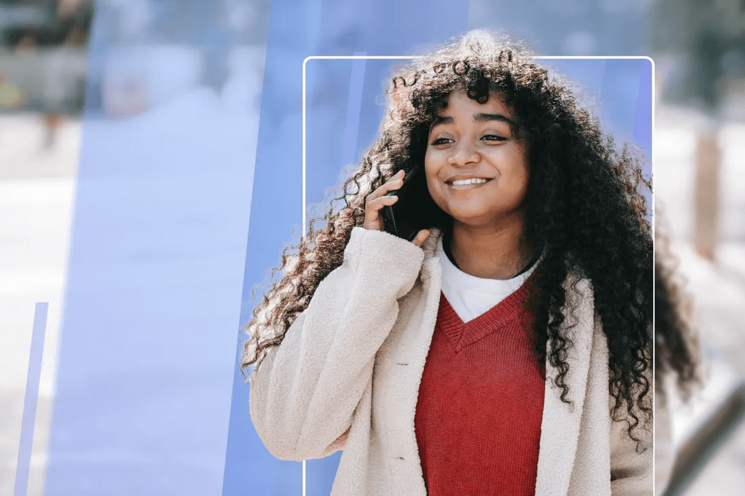 Young woman with long curly hair smiling warmly, wearing a red sweater and beige cardigan talking on mobile phone.