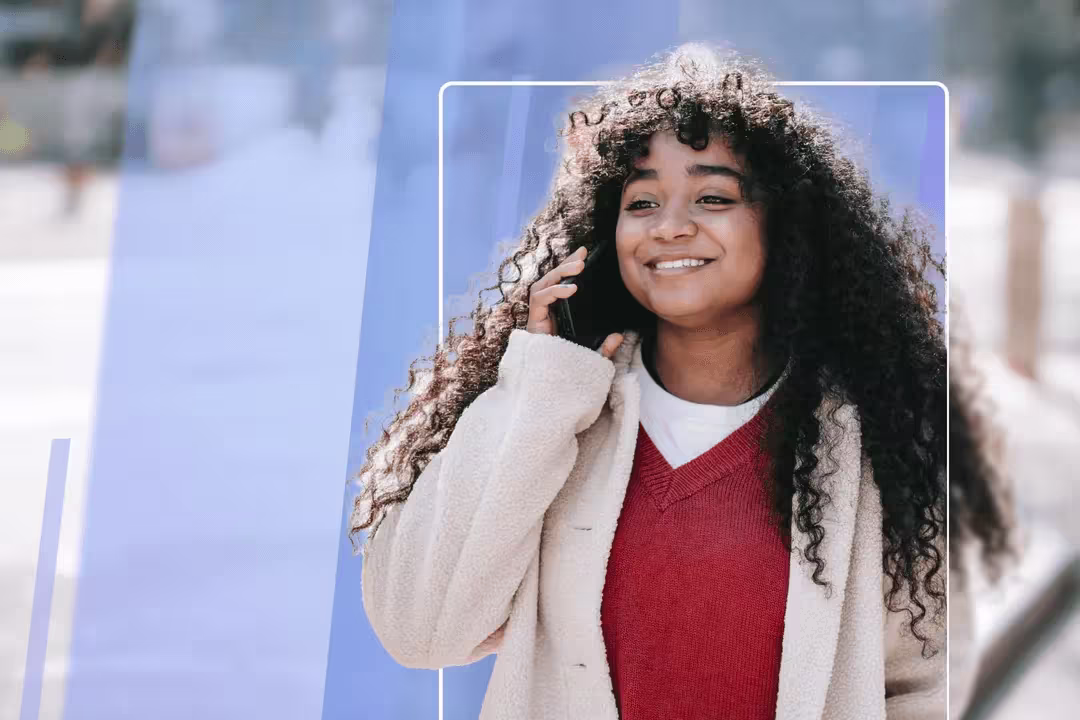 Young woman with long curly hair smiling warmly, wearing a red sweater and beige cardigan talking on mobile phone.