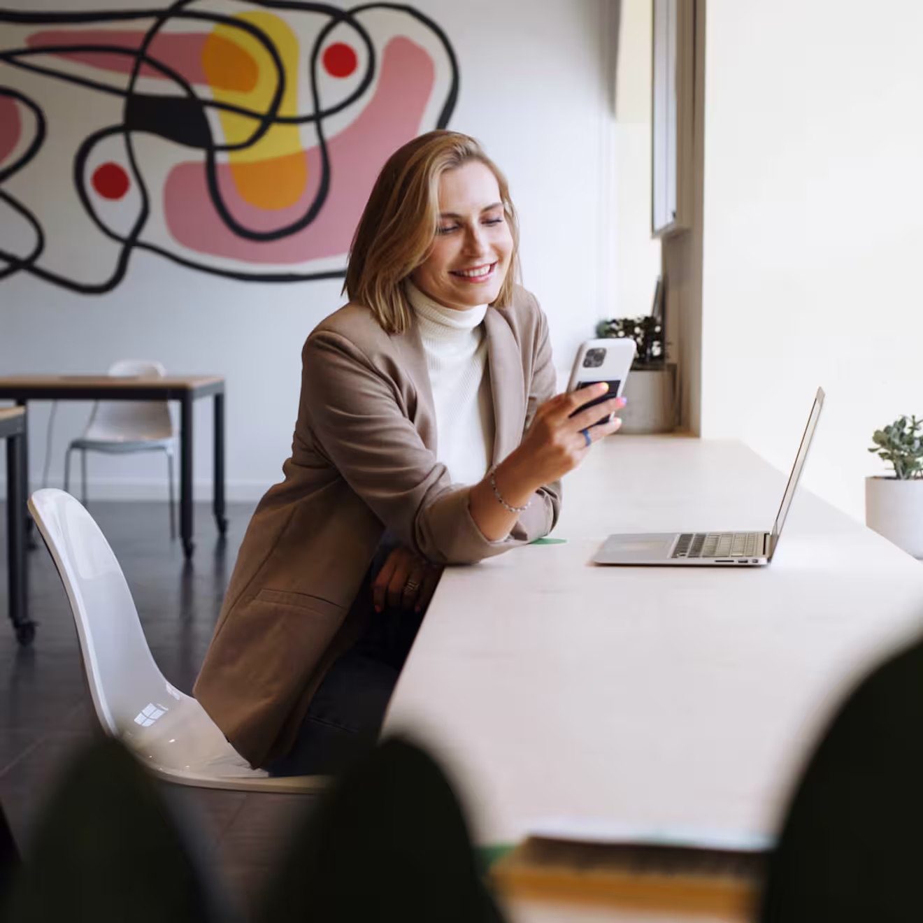 Smiling woman in tan blazer uses smartphone at desk with laptop in bright modern office with colorful mural