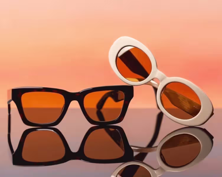 Two pairs of retro sunglasses with amber lenses reflected on glossy surface against coral backdrop