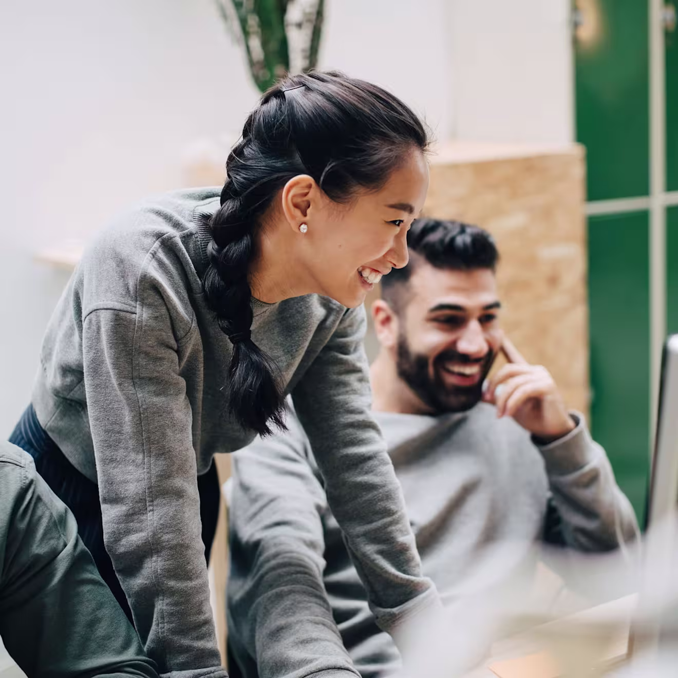 Two young professionals in casual grey sweaters share a genuine laugh while collaborating in a bright office space
