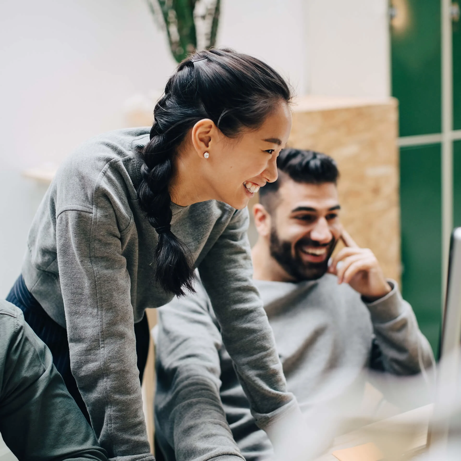 Two young professionals in casual grey sweaters share a genuine laugh while collaborating in a bright office space