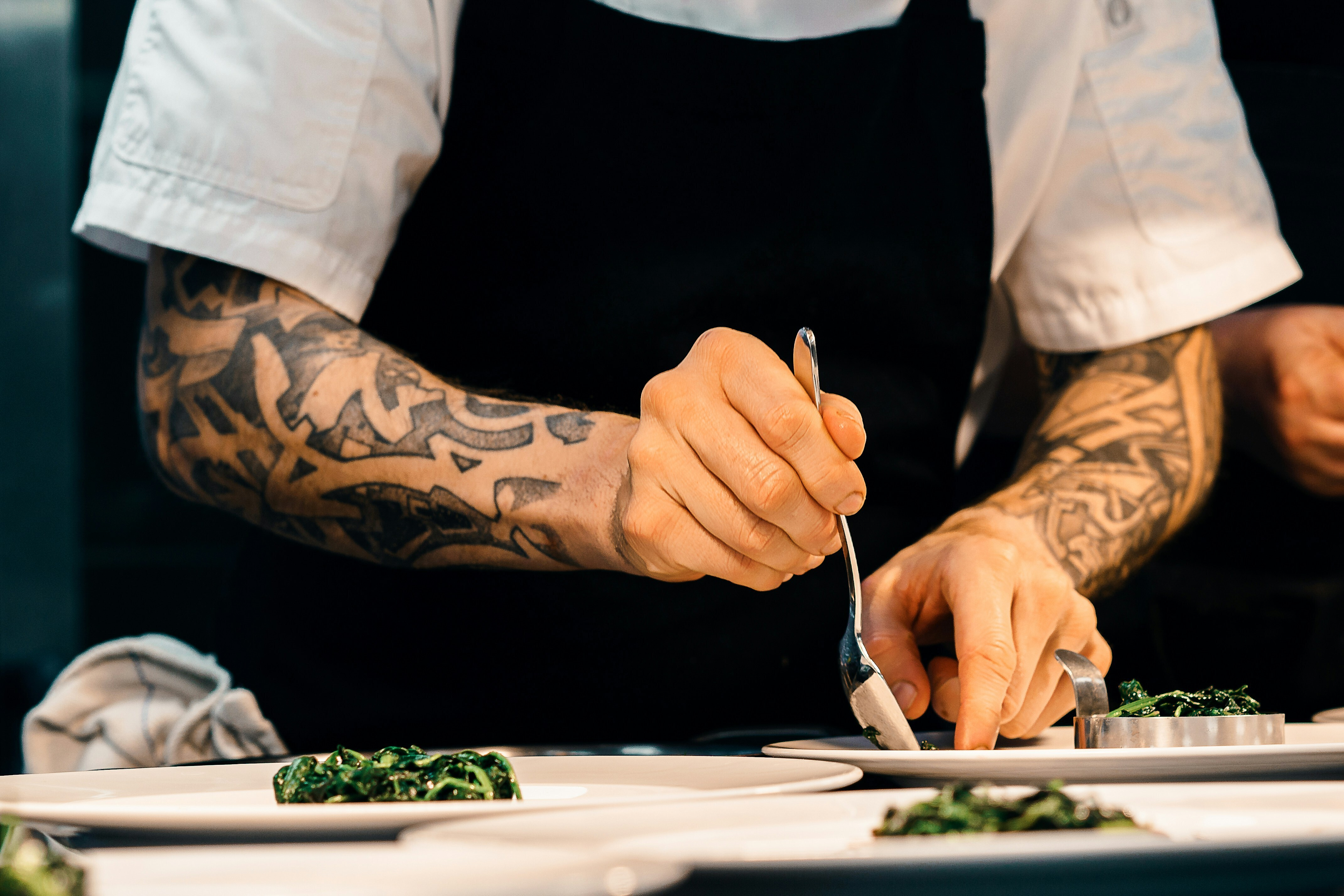 Tattooed chef in black apron and white sleeves plating green vegetables on white dishes in a professional kitchen