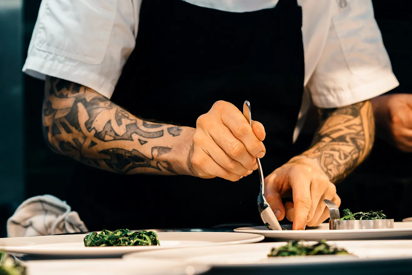 Tattooed chef in black apron and white sleeves plating green vegetables on white dishes in a professional kitchen