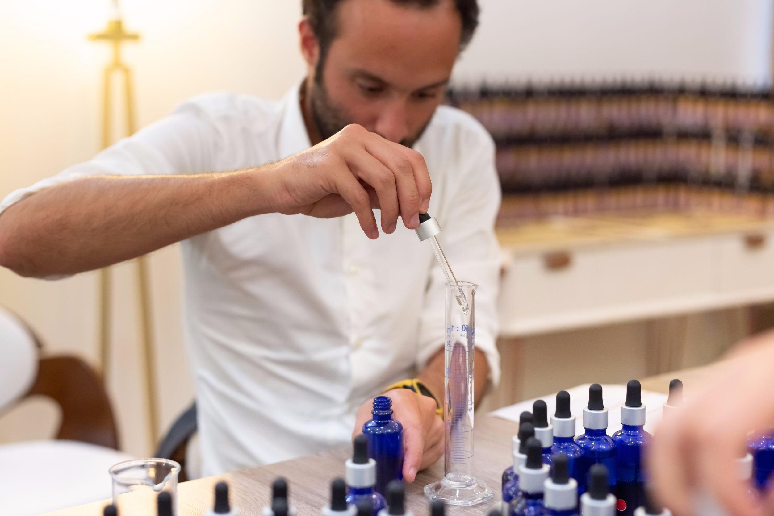 Perfumer in white shirt carefully measures liquid ingredients using dropper and graduated cylinder, surrounded by blue glass bottles