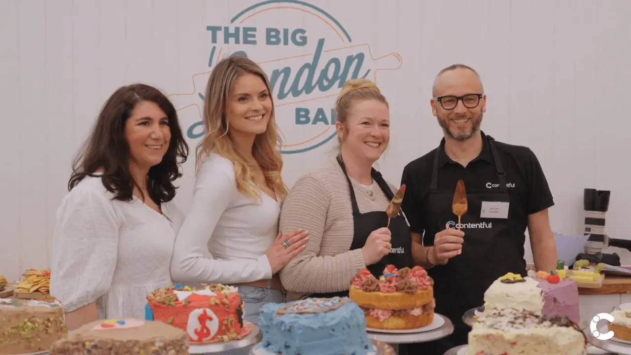 Group of smiling people standing behind a table displaying colorful decorated cakes at "The Big London Bake" event