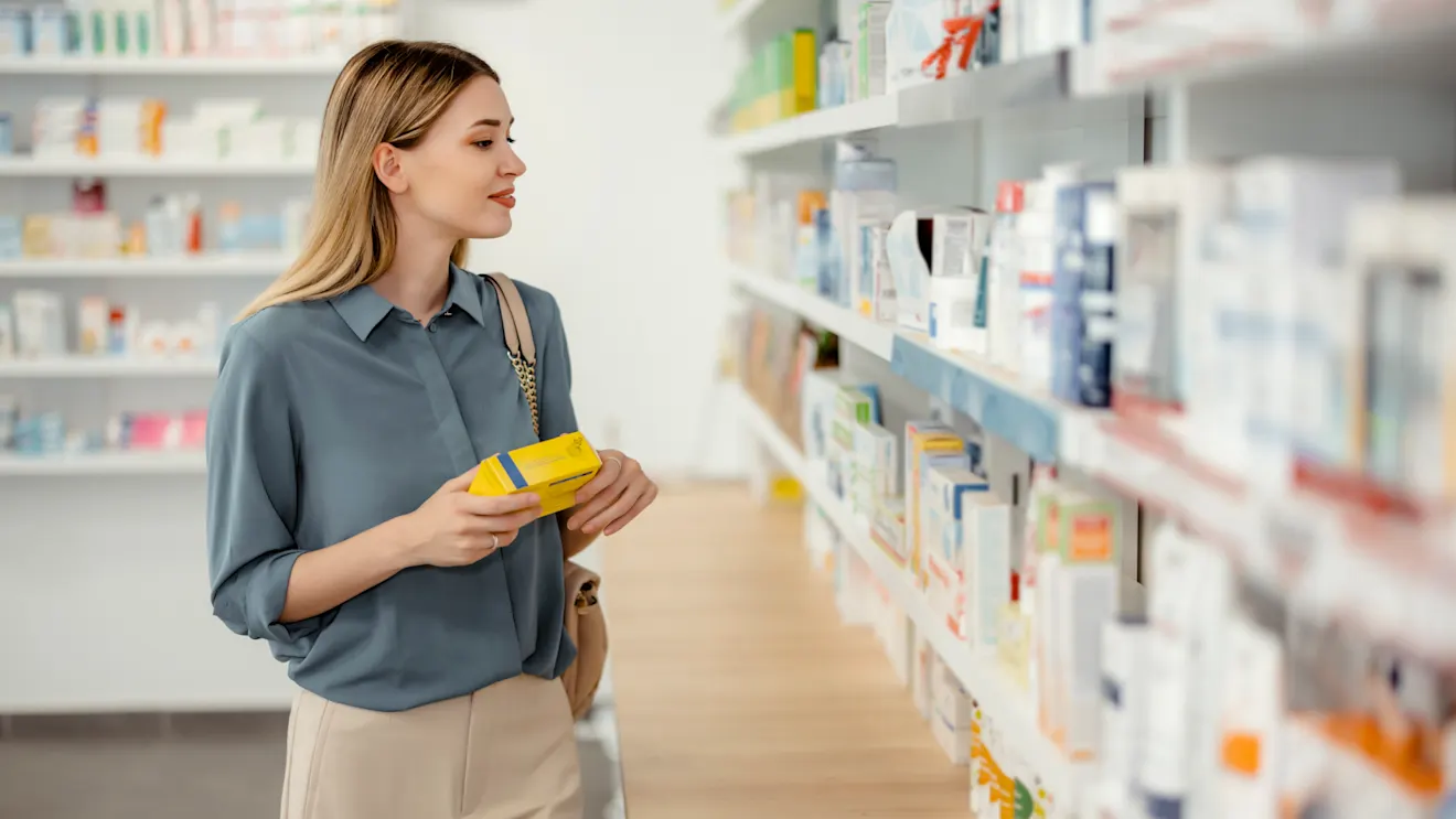 A young blonde woman in a blue blouse holds a yellow product box while browsing shelves in a pharmacy.