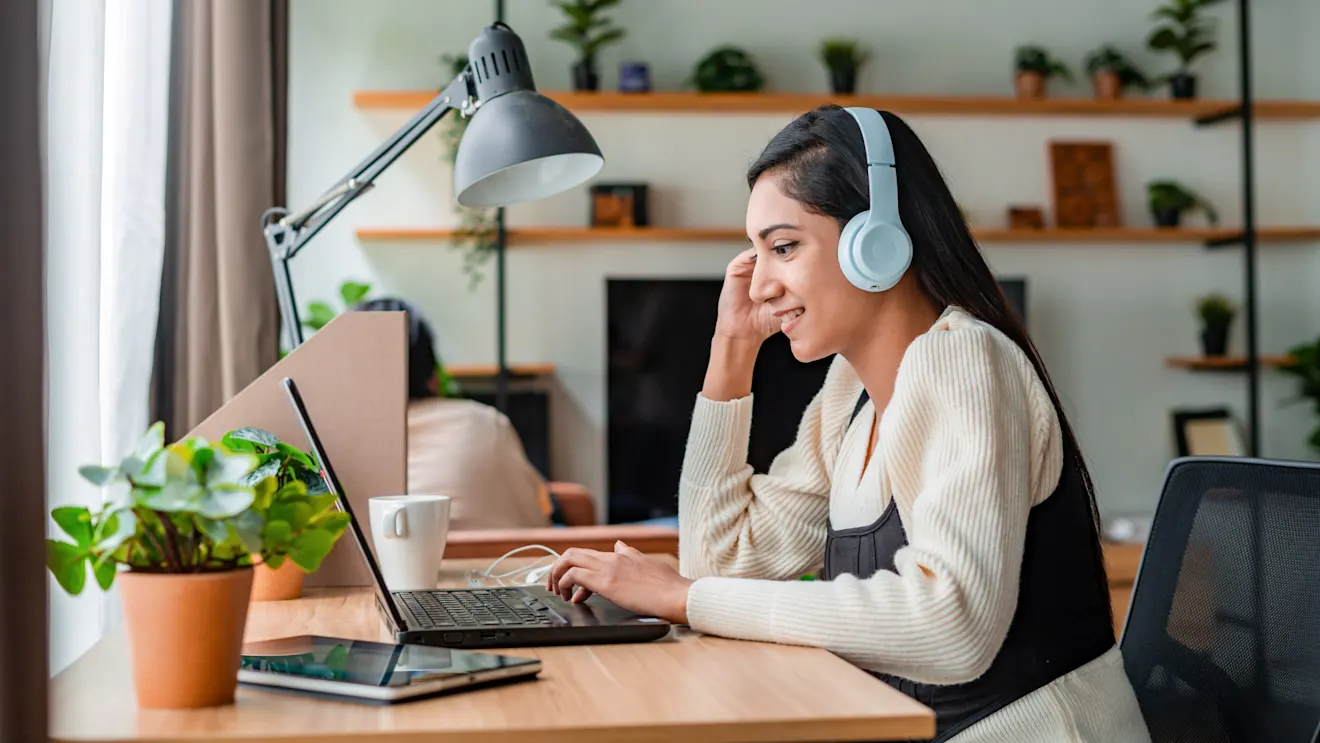 A smiling young woman wearing white headphones works on a laptop at a wooden desk in a cozy home office with plants.