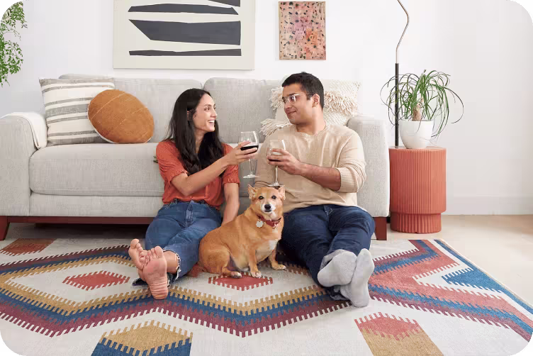 Couple relaxing on patterned rug with wine glasses and their ginger-colored dog, modern living room with abstract wall art