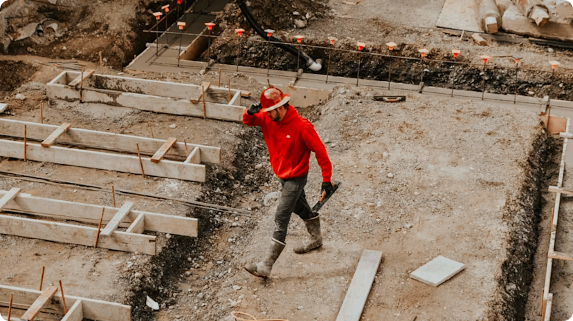 Man in a red hard hat, red hoodie, black trousers and black boots walking across a construction site