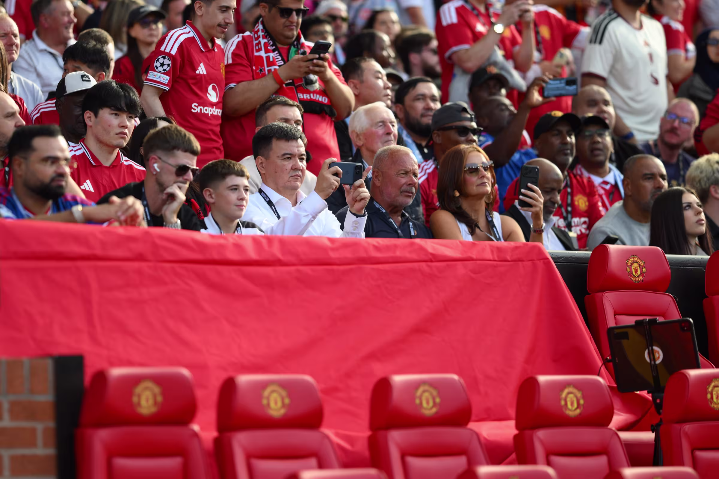 Rows of empty red Manchester United crested seats beside the dugout, with supporters filling the stand behind them before kick-off.
