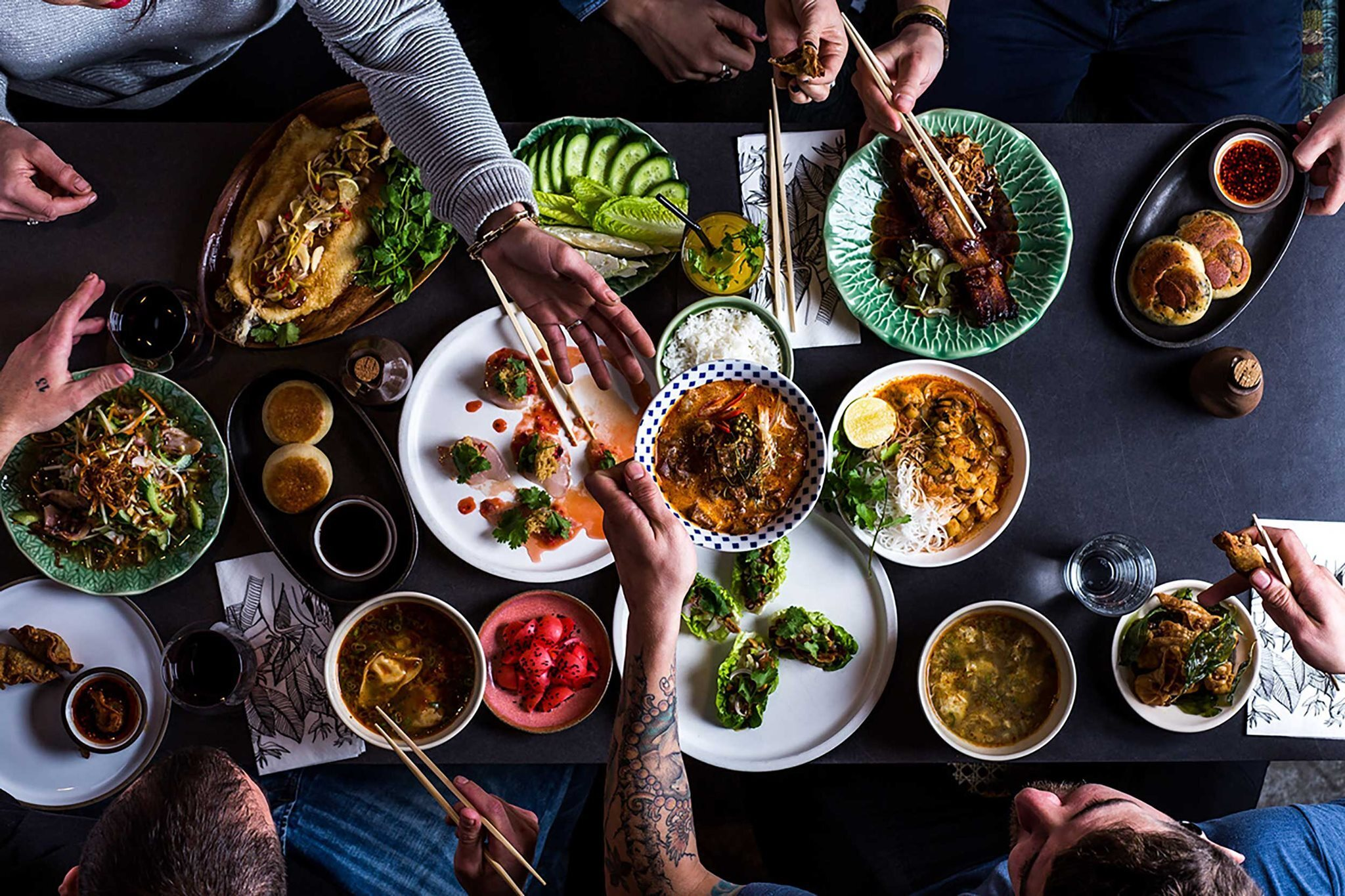 Overhead view of Asian feast with various dishes, soups, and sides on dark table, with hands holding chopsticks reaching for food
