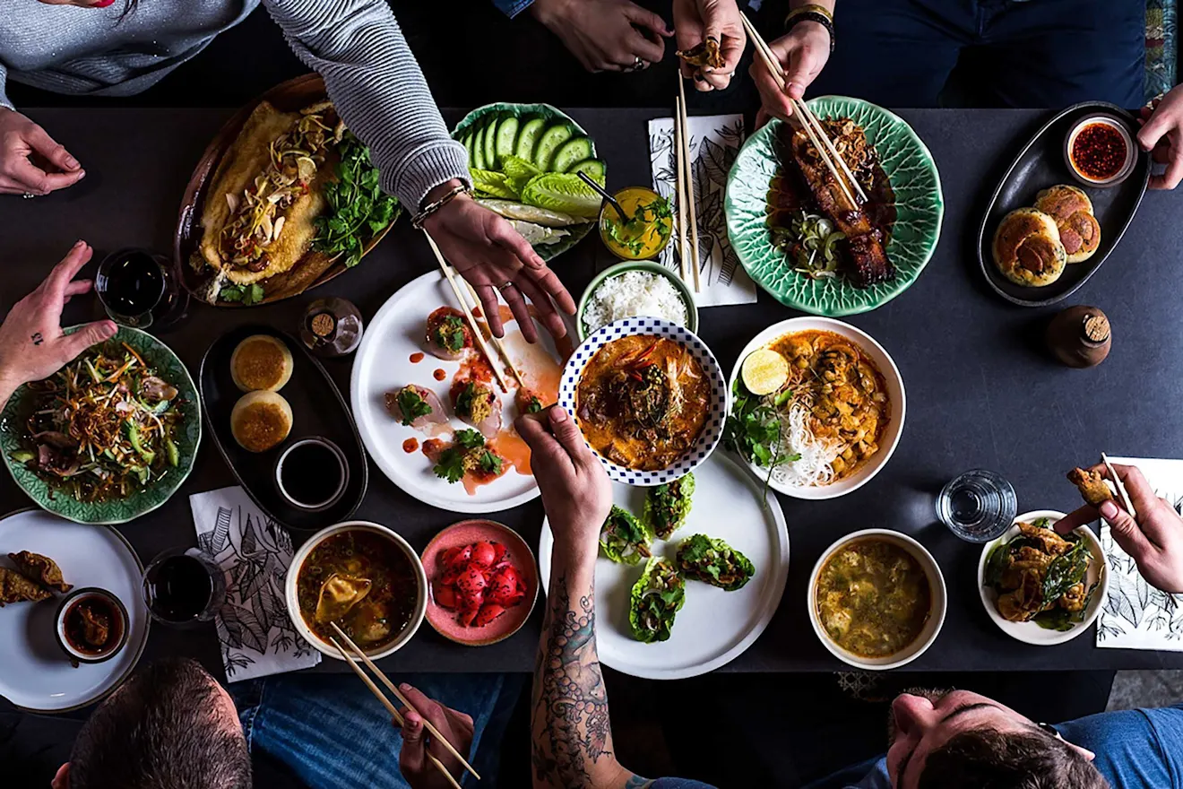 Overhead view of Asian feast with various dishes, soups, and sides on dark table, with hands holding chopsticks reaching for food