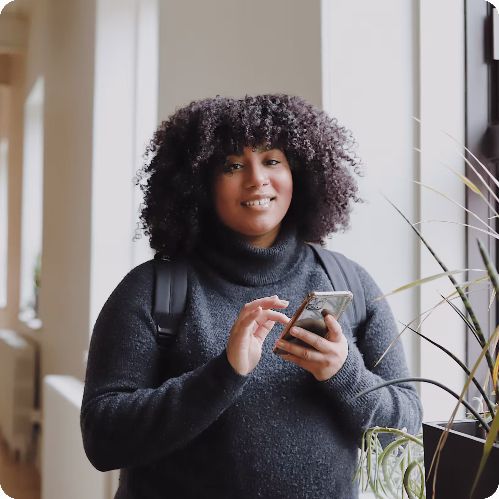 Woman in black turtleneck smiles while holding gold phone in bright office corridor near large window and plant.