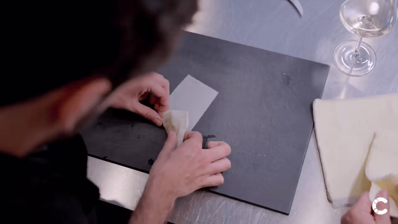 Hands folding pasta on a dark surface with wine glasses visible in the background
