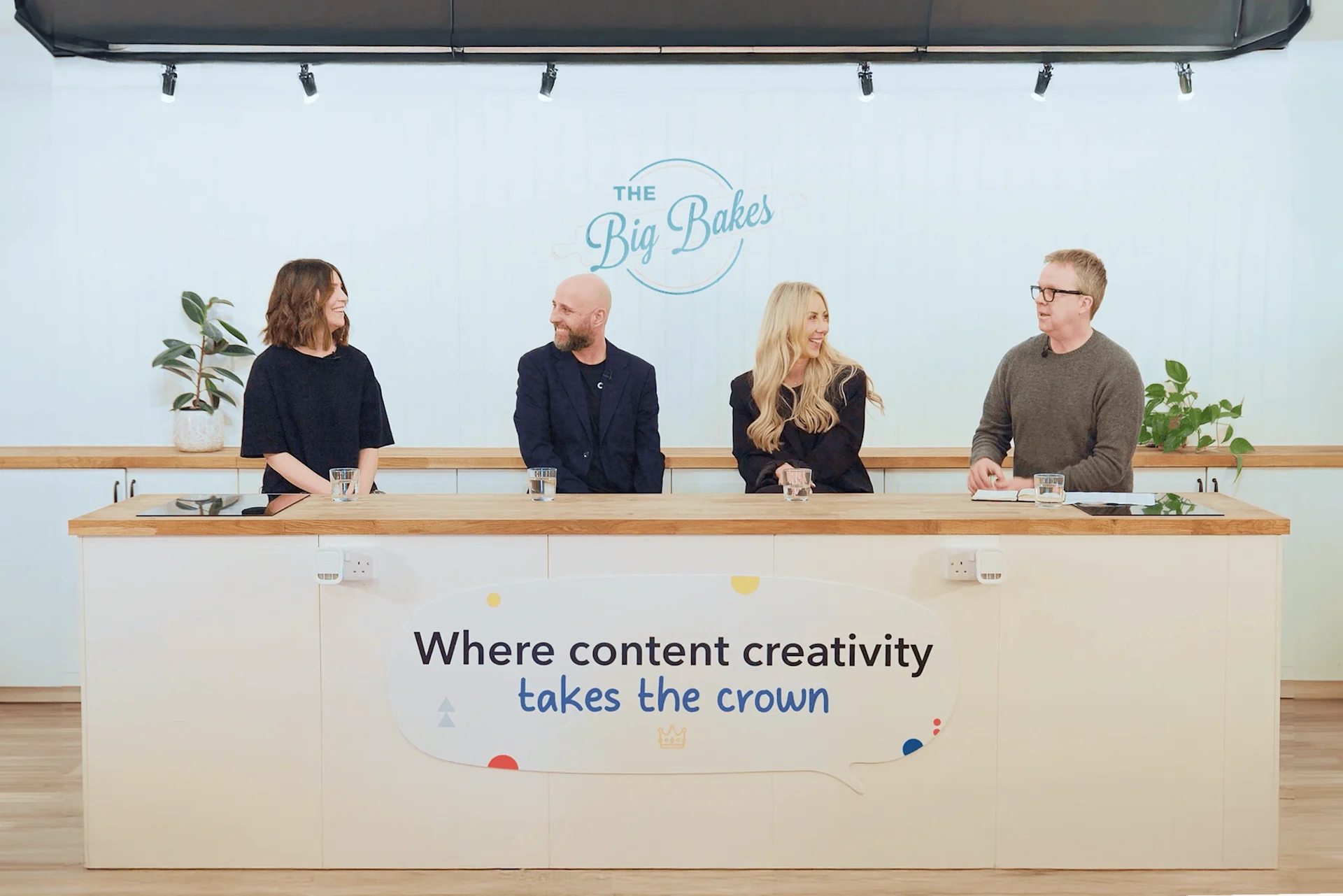 Four team members seated at a desk with "The Big Bakes" logo and text "Where content creativity takes the crown" on wall behind them