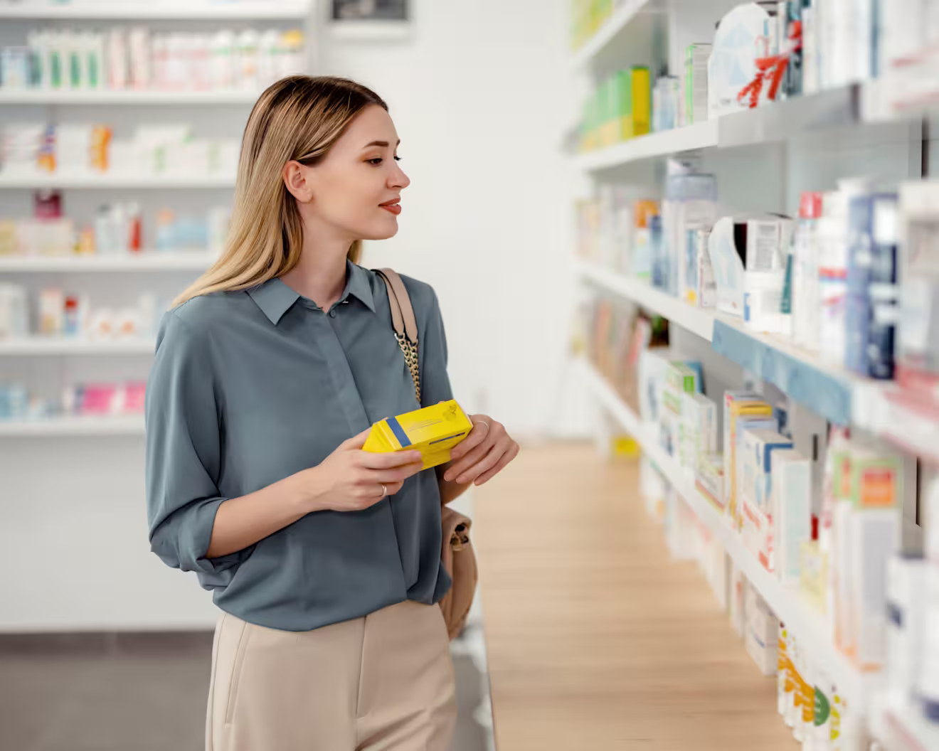 A young blonde woman in a blue blouse holds a yellow product box while browsing shelves of medications in a pharmacy.