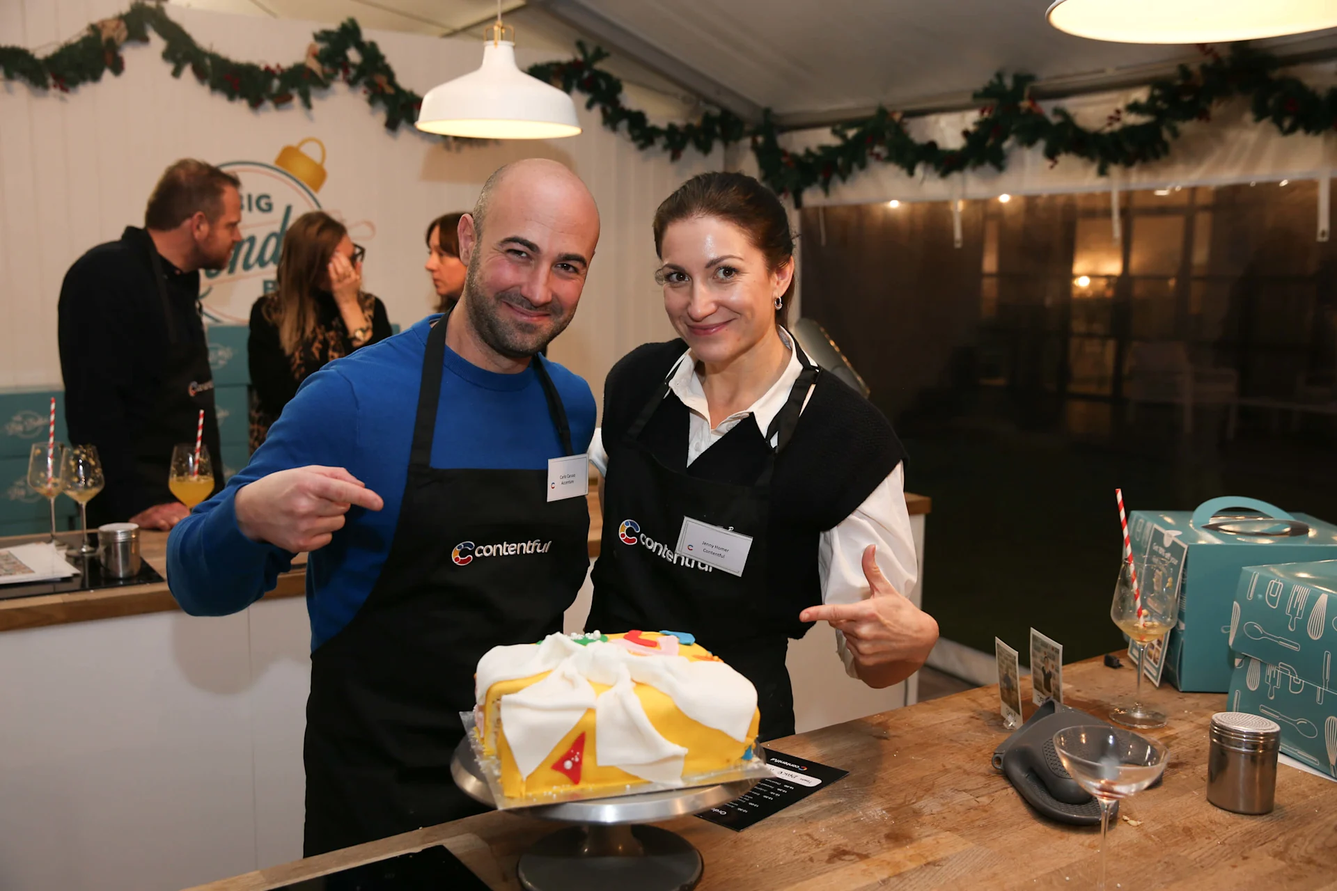 Two Contentful team members in black aprons present a decorated white and yellow cake at a holiday event with evergreen garlands