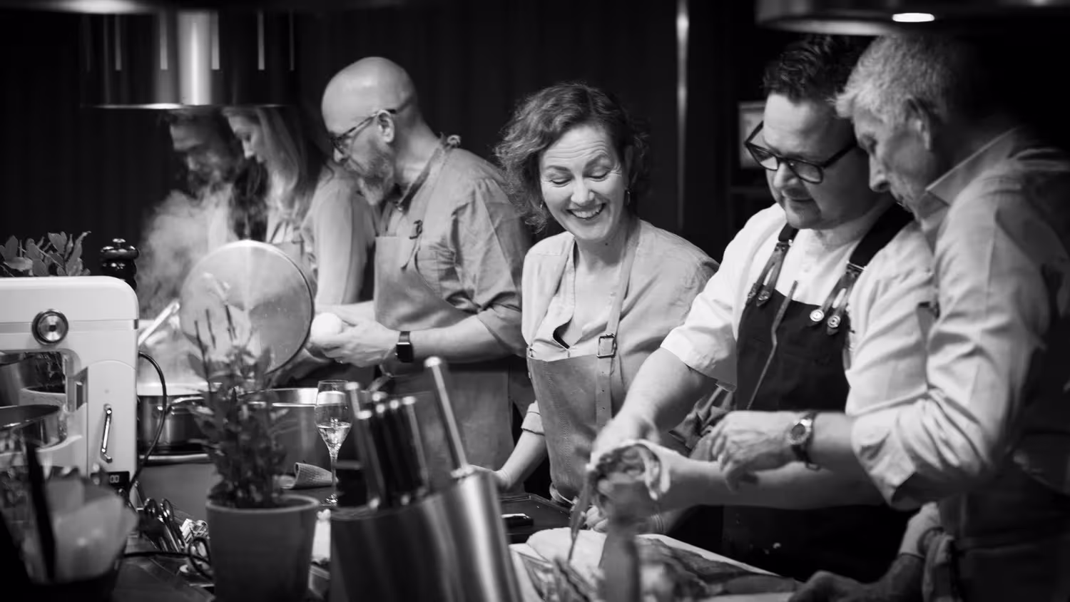 Black and white photo of people in aprons preparing food together in a professional kitchen.