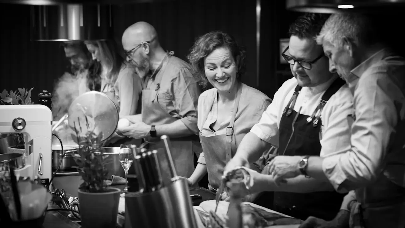 Black and white photo of people in aprons preparing food together in a professional kitchen.