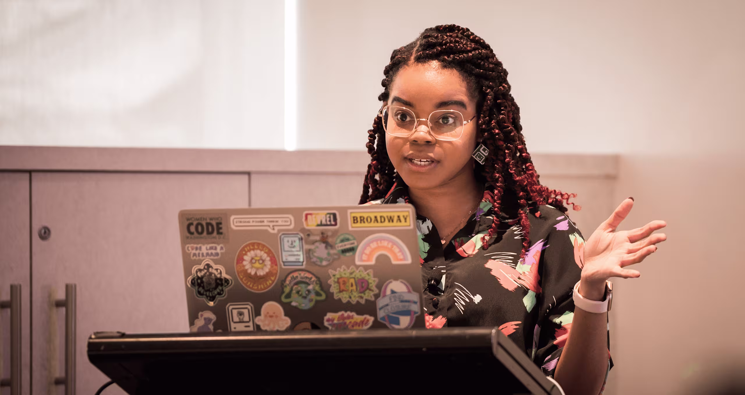 Professional speaker with braided hair and glasses gestures while presenting, laptop covered in coding and tech-related stickers visible