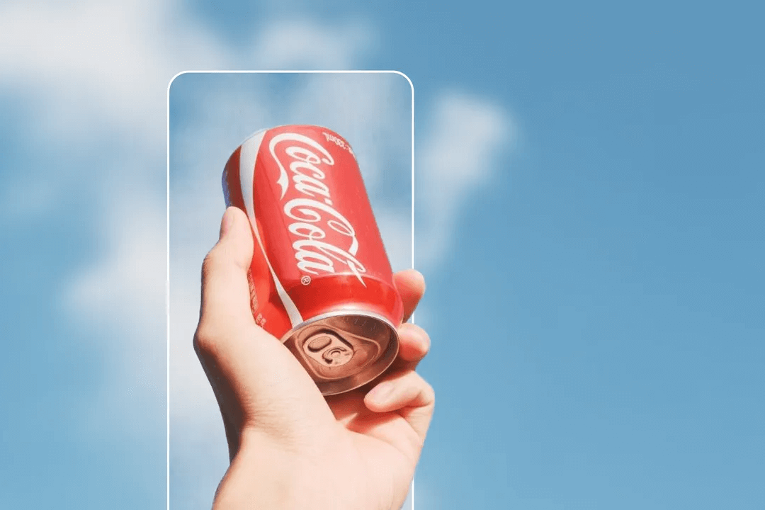 Hand holding a red Coca-Cola can against a bright blue sky with white clouds