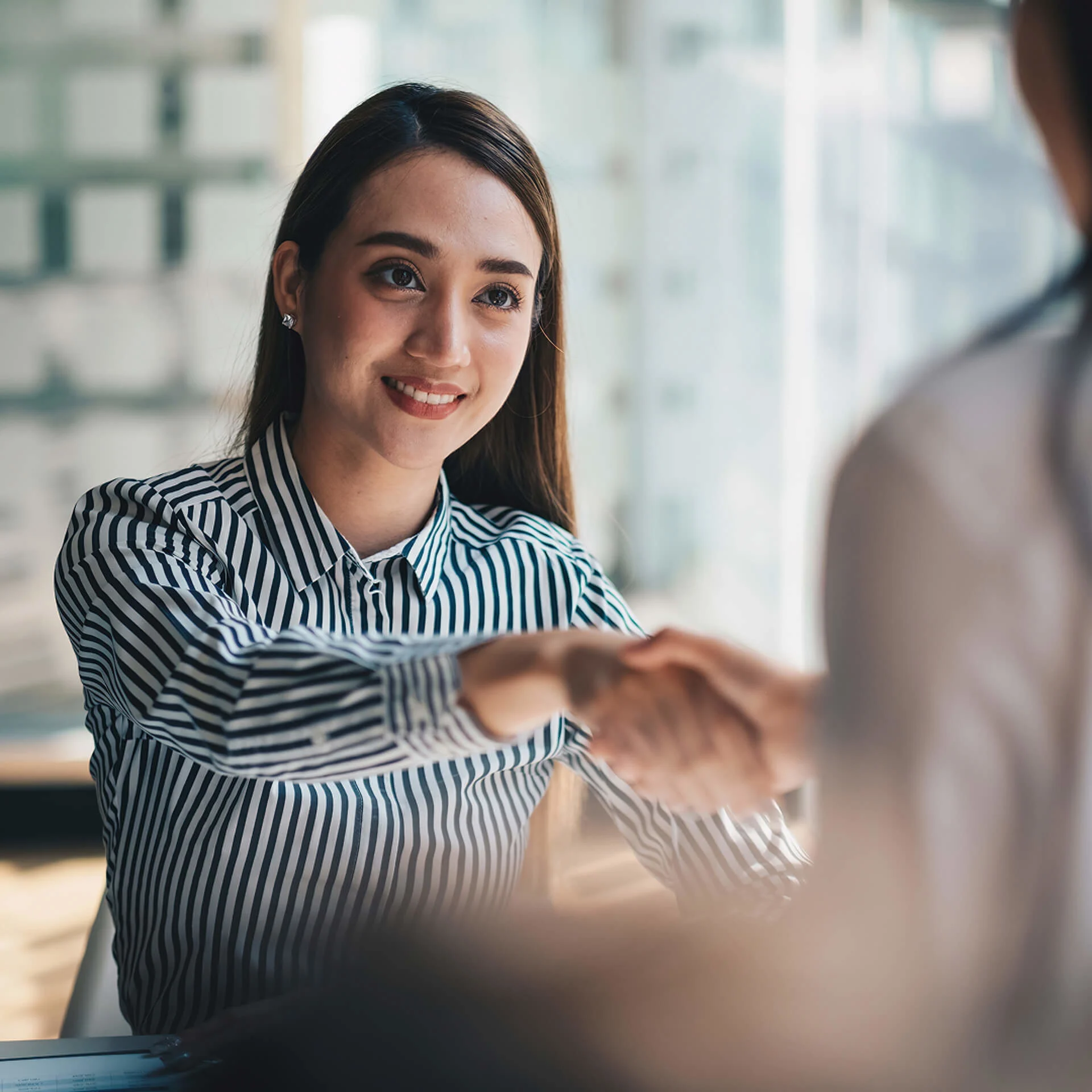 Professional in striped blouse shaking hands with colleague during business meeting in bright office setting