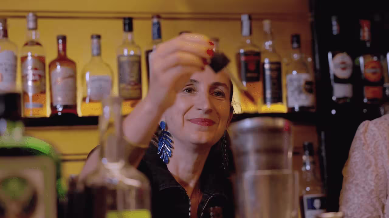 Bartender in front of illuminated liquor bottles on yellow wall, wearing blue statement earrings and smiling warmly