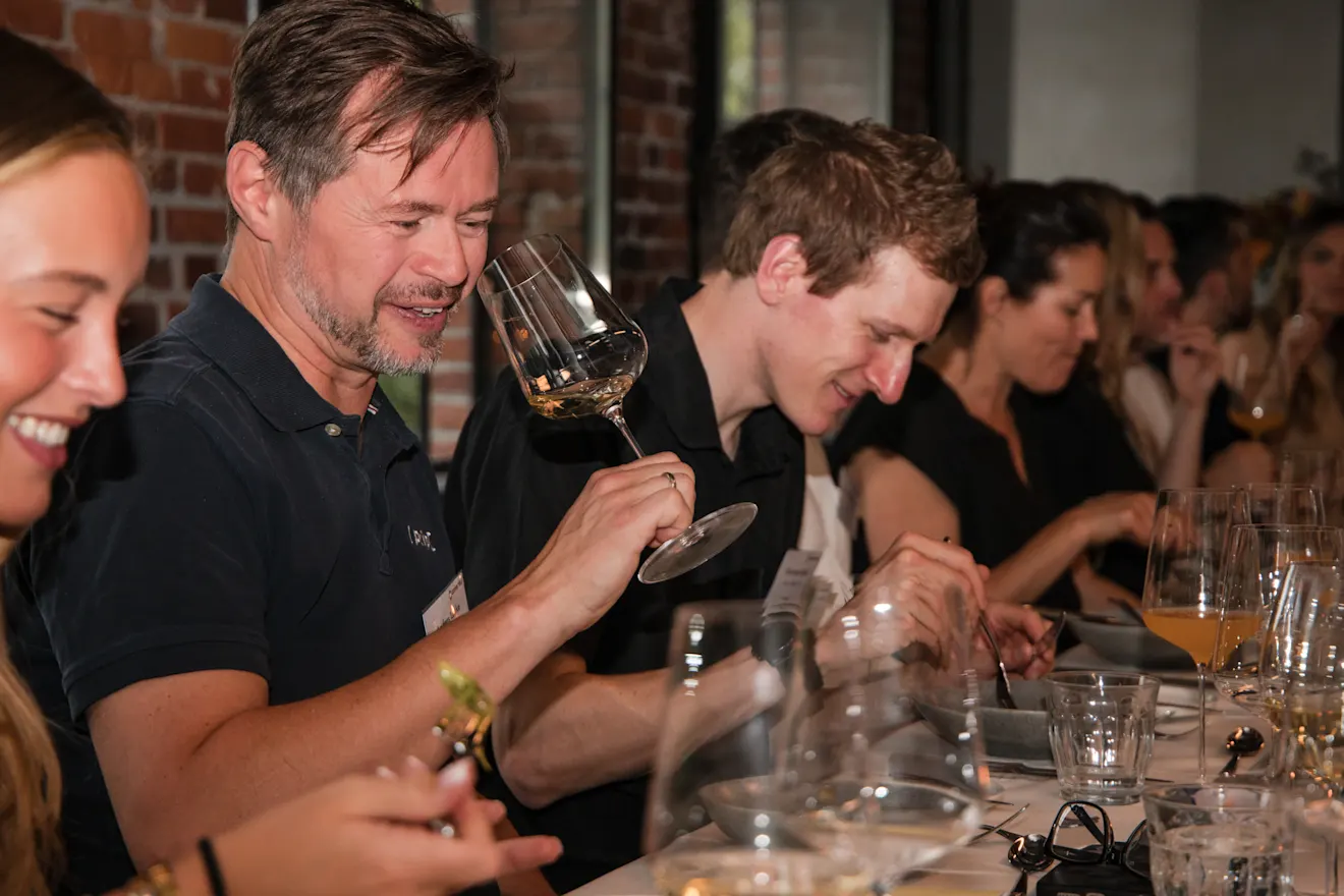 Group dining at a long table with wine glasses, with a smiling man in a navy shirt holding up a glass.