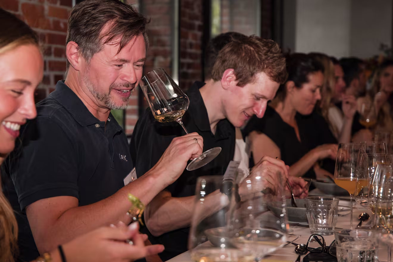 Group dining at a long table with wine glasses, with a smiling man in a navy shirt holding up a glass.